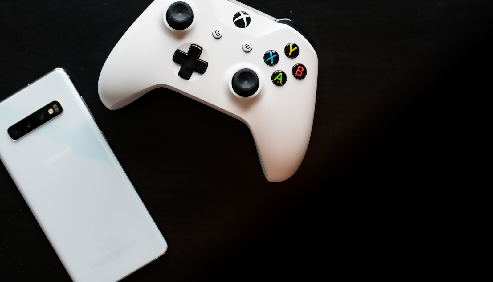 Shot taken from above against a dark wooden background showing a White Samsung Galaxy S10+ and a Xbox One White Controller next to each other with room for text on the right hand side